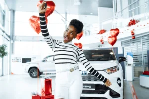 Mulher sorridente, usando blusa listrada branca e preta e calça branca, segurando a chave de um carro novo em uma concessionária decorada com balões vermelhos em forma de coração e presentes ao fundo. Um carro branco está estacionado atrás dela.