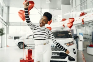 Mulher sorridente, usando blusa listrada branca e preta e calça branca, segurando a chave de um carro novo em uma concessionária decorada com balões vermelhos em forma de coração e presentes ao fundo. Um carro branco está estacionado atrás dela.