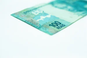 Nota de dinheiro de 100 reais parcialmente visível sobre uma superfície branca. A cédula tem tonalidade azul-esverdeada, com detalhes gráficos característicos da moeda brasileira. O número "100" e a palavra "REAIS" estão em destaque no canto inferior direito.