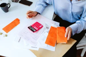 Pessoa vestindo camisa azul analisando recibos e documentos fiscais em uma mesa branca, com uma calculadora rosa ao lado. A imagem representa a organização de despesas e impostos.