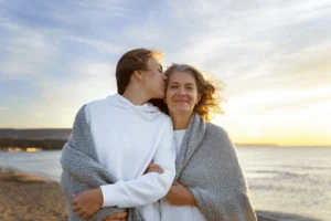 Duas mulheres abraçadas na praia ao pôr do sol, com uma delas beijando a outra na bochecha e ambas sorrindo.