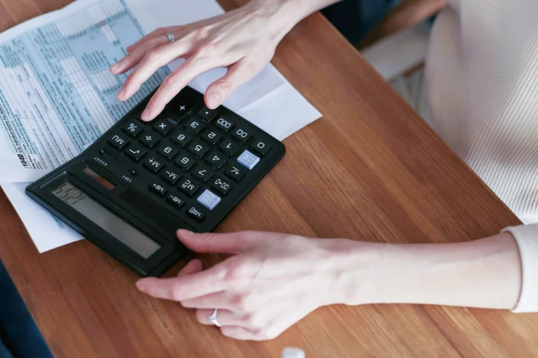 Pessoa usando uma calculadora sobre uma mesa de madeira, com papéis e formulário ao fundo.