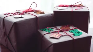 Três caixas de presente embrulhadas em papel pardo, amarradas com barbante vermelho e decoradas com pequenos enfeites natalinos de feltro, como árvore, meia e bonequinho de gengibre.