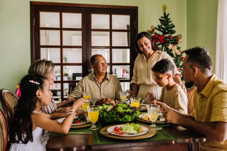 Família reunida à mesa para a ceia de Natal, com pratos de comida, bebidas e árvore de Natal decorada ao fundo