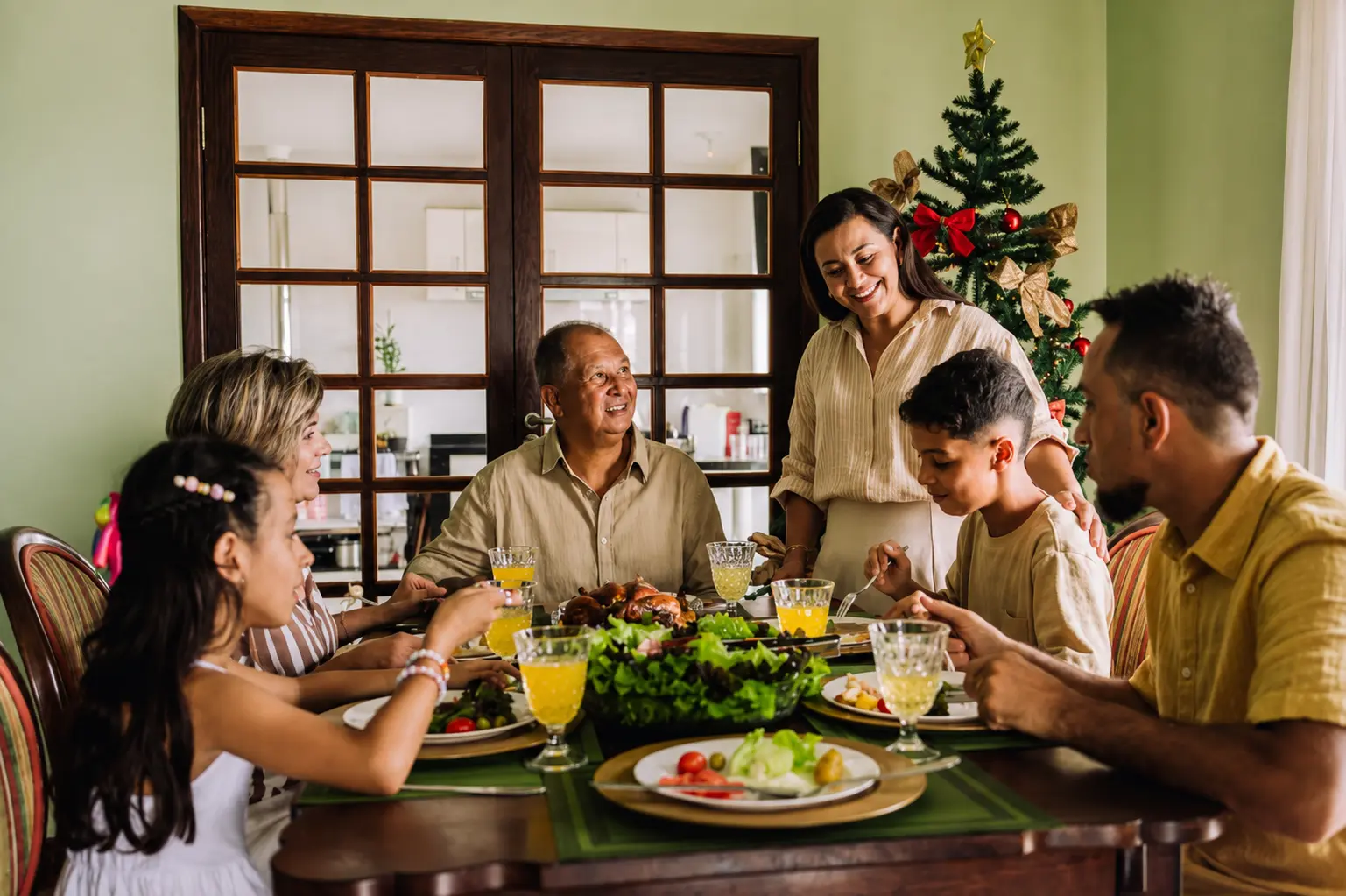 Família reunida à mesa para a ceia de Natal, com pratos de comida, bebidas e árvore de Natal decorada ao fundo